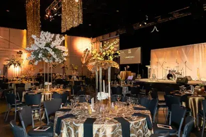 Elegant gold‑themed gala event — round tables with black chairs, tall floral centerpieces, string lights and draped fabric overhead in a large banquet hall.