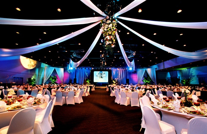 Large, elegant corporate event venue: round tables with white tablecloths and chairs, soft lighting, draped fabric hanging in a star pattern from the ceiling, and a floral centerpiece overhead.
