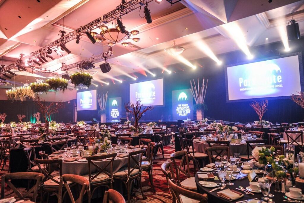 Elegant gala event — people dressed in formal attire seated at round tables under soft lighting, with a stage or podium in the background.