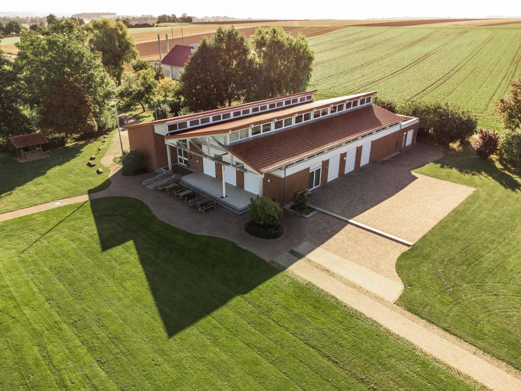 Aerial view of Bürgerhaus Krebeck — a red-brick community building with a sloped roof, set in a green, tree-lined landscape.